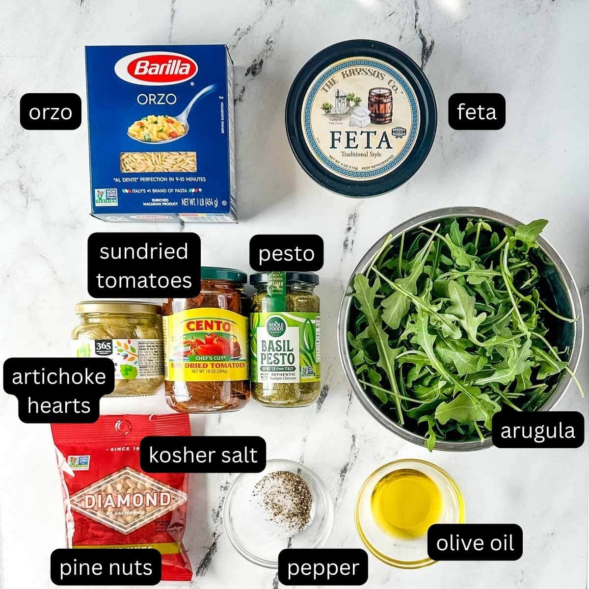 The labeled ingredients for orzo pesto salad sit on a white marble counter.