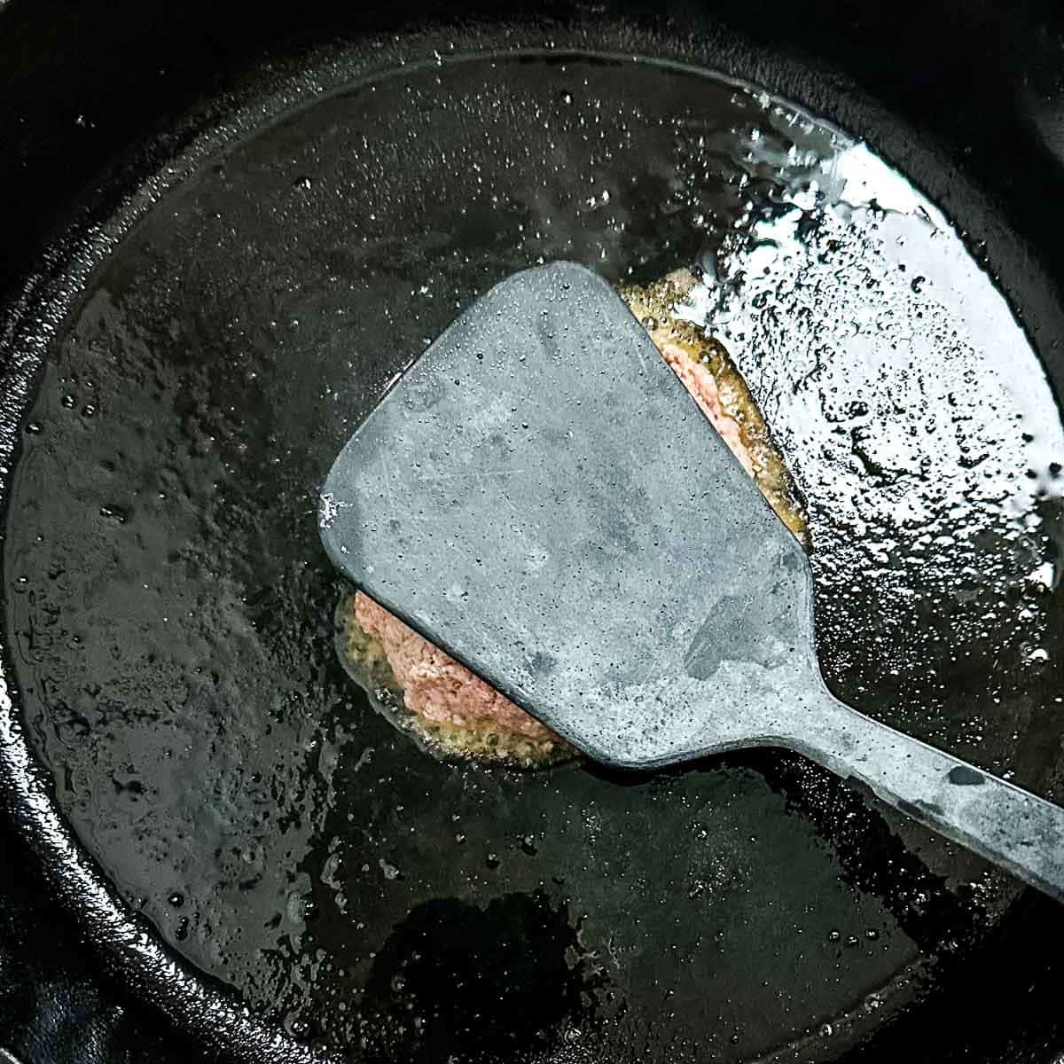 turkey burger patty is smashed with a black spatula in a cast iron pan.