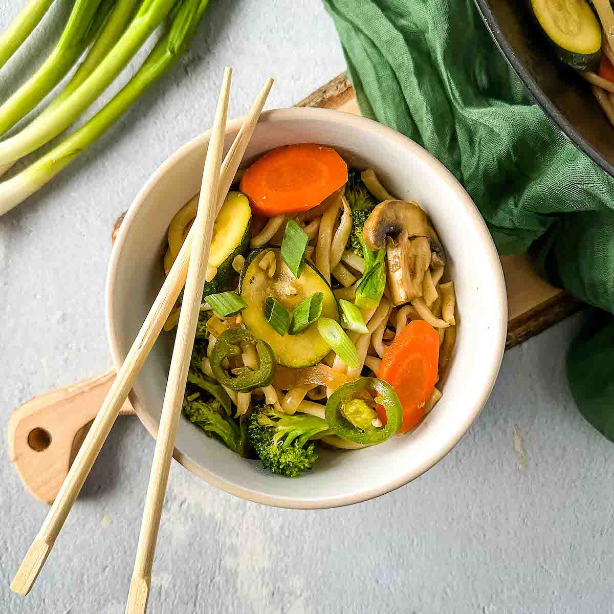 A bowl of vegetable yaki udon is shown with wooden chopsticks, a wok of yaki udon, and scallions in the background.