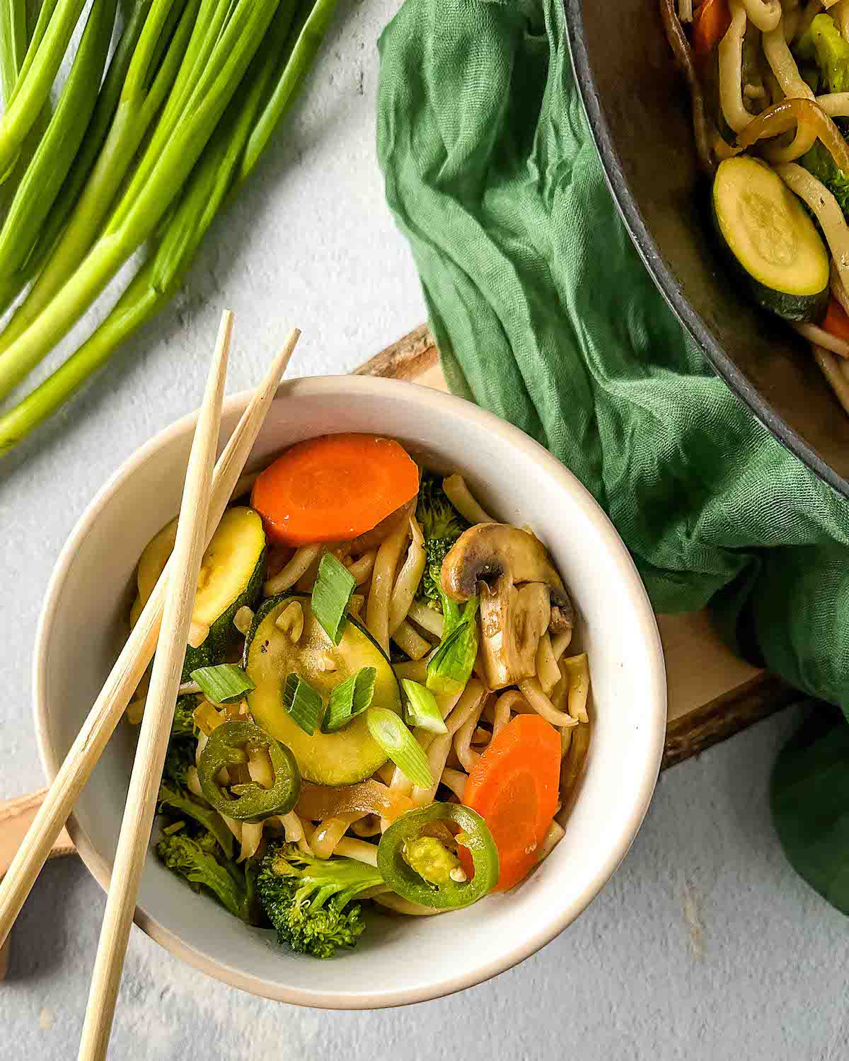 A bowl of vegetable yaki udon is shown with wooden chopsticks, a wok of yaki udon, and scallions in the background.