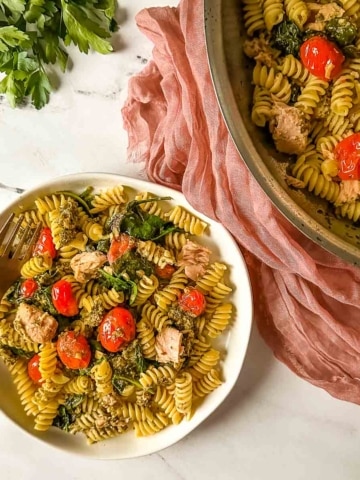 A plate of tuna pesto pasta with cherry tomatoes, a copper fork, and a pink linen next to a pan full of tuna pesto pasta.