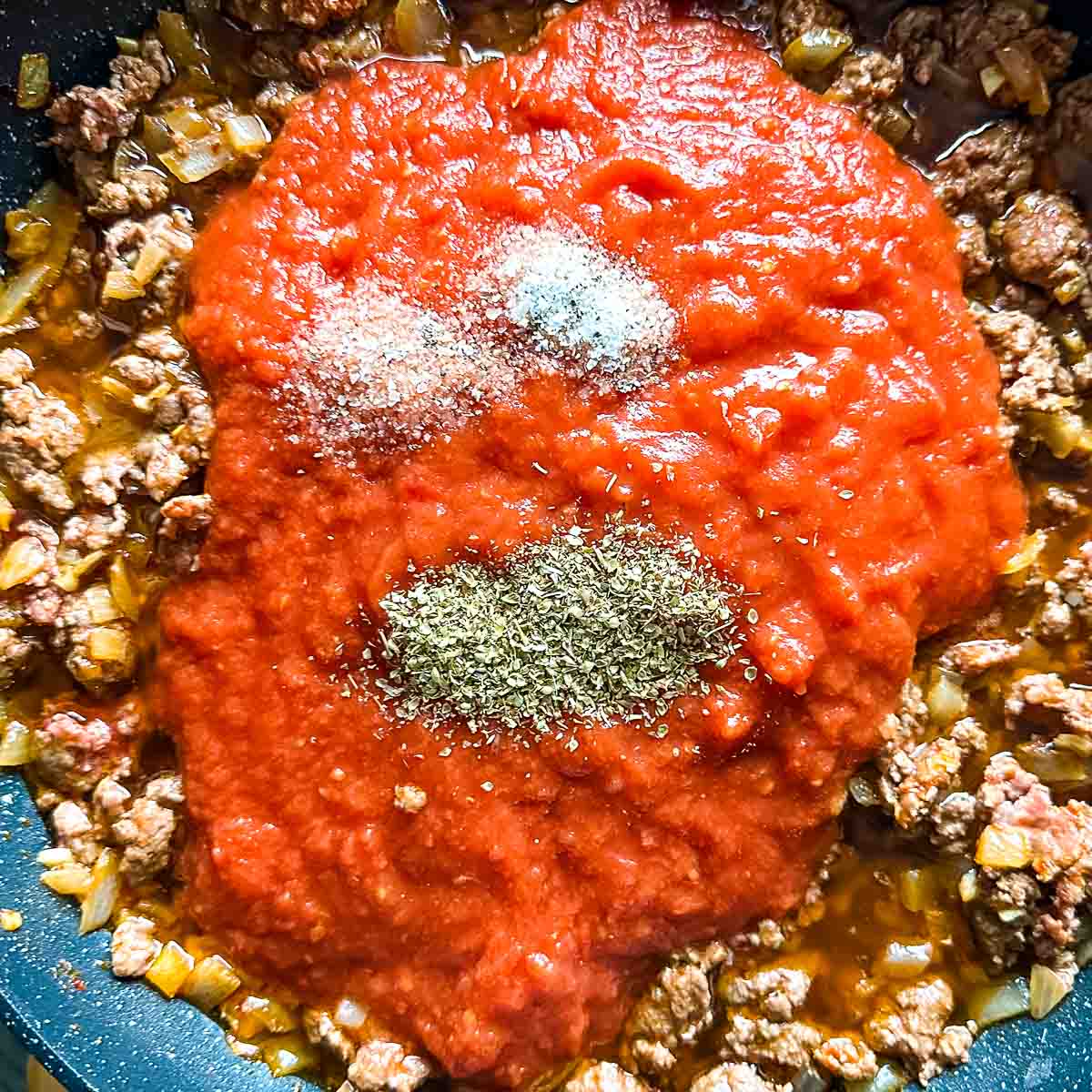 Crushed tomatoes, salt, and Italian spices are added to the lamb mixture in the frying pan.