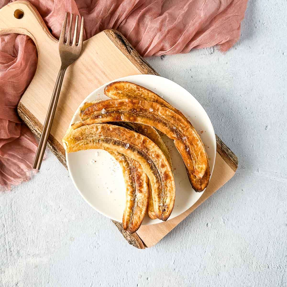 air fryer bananas on a white plate over a rustic wooden cutting board with a pink linen and a copper fork.