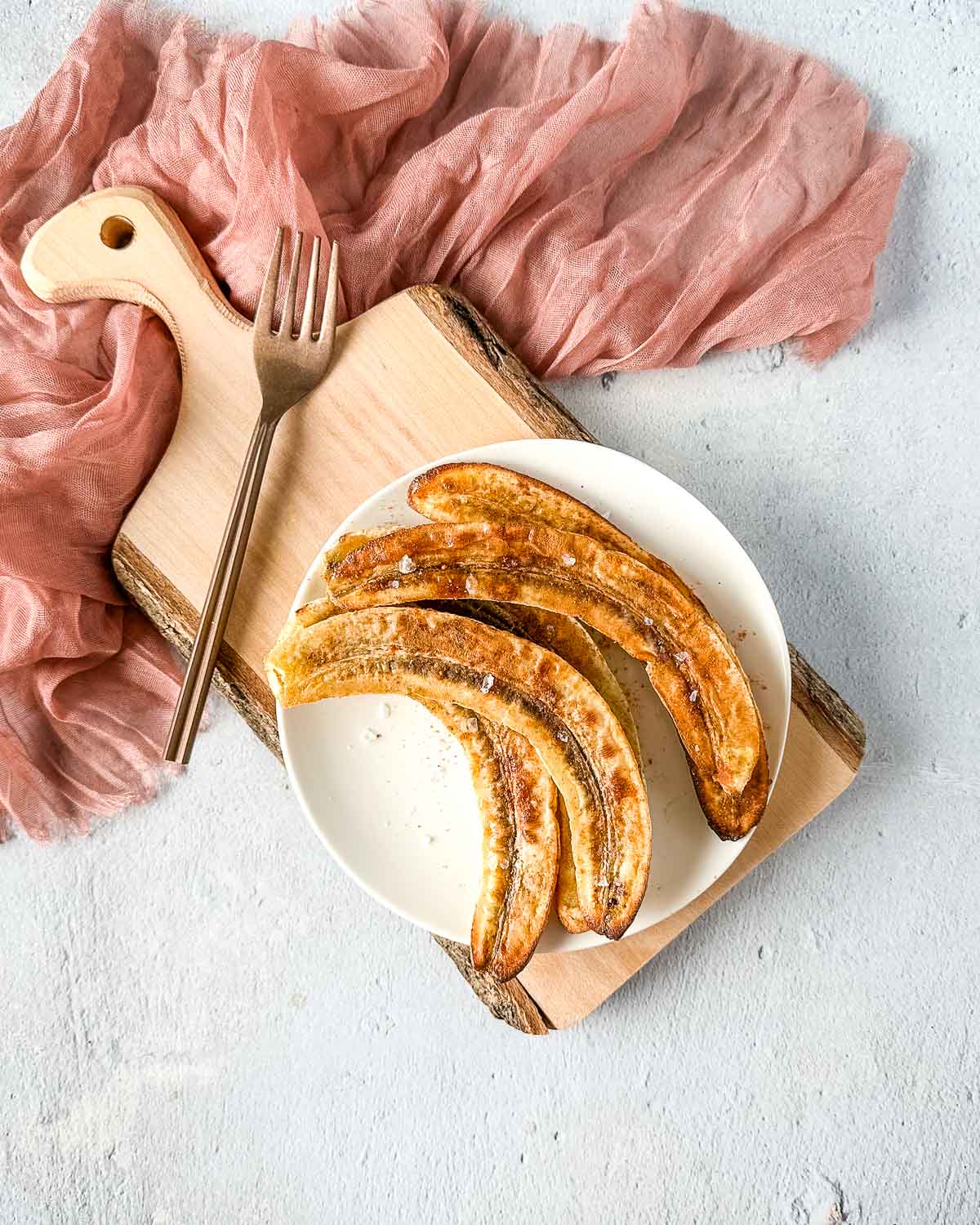 air fryer bananas on a white plate over a rustic wooden cutting board with a pink linen and a copper fork.