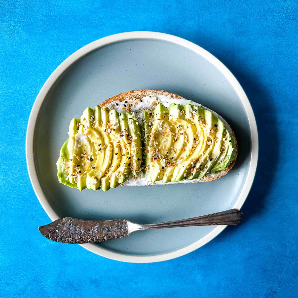 an avocado tartine sits on a light blue plate with a vintage silver knife over a blue darker background.