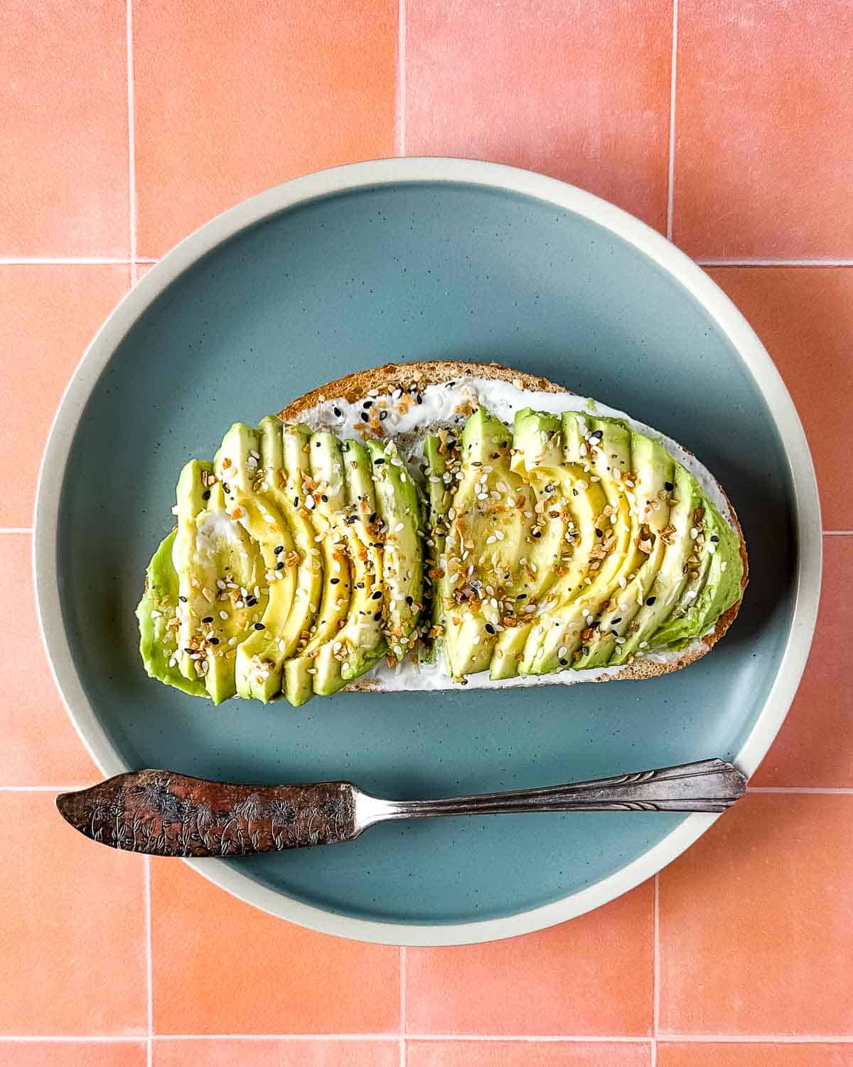 an avocado tartine sits on a blue plate with a vintage silver knife over a peach tile background.
