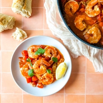 plate of bloody mary shrimp next to a pan of shrimp with a piece of baguette and a white linen.