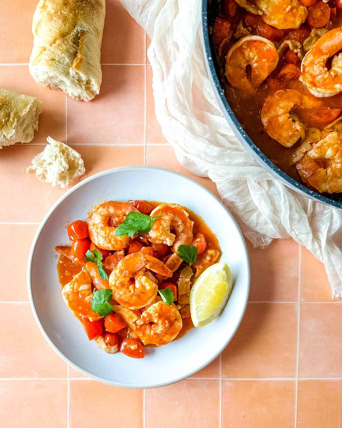 plate of bloody mary shrimp next to a pan of shrimp with a piece of baguette and a white linen.