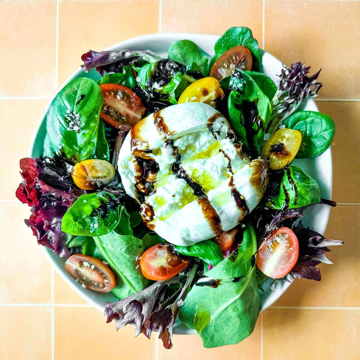 burrata salad with balsamic glaze including spring mix, basil leaves, and halved cherry tomatoes drizzled with balsamic glaze and olive oil.