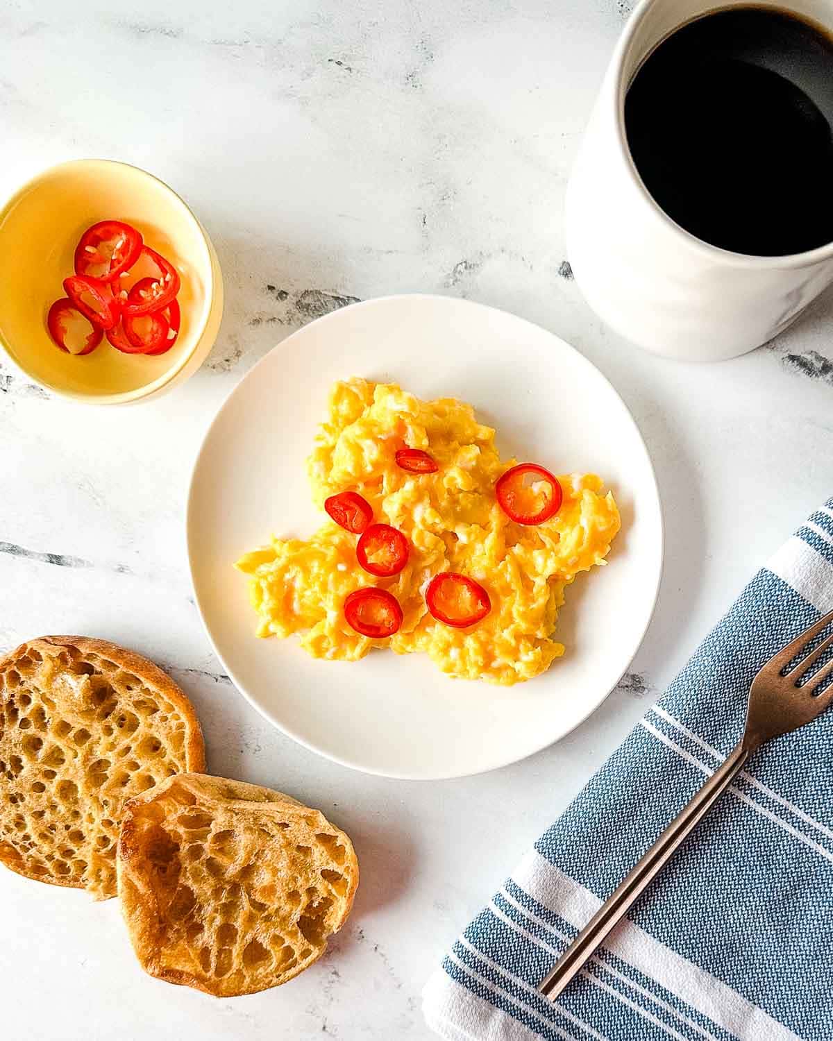 chilli scrambled eggs on a white plate surrounded by a cup of coffee, English muffins, and a copper fork.
