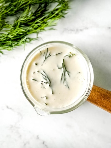 closeup of dill sauce in a glass container with a wooden handle on a white marble counter.