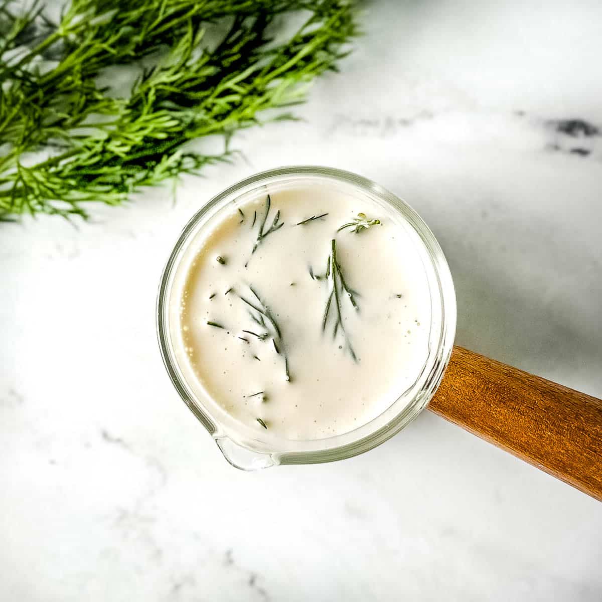 closeup of dill sauce in a glass container with a wooden handle on a white marble counter.