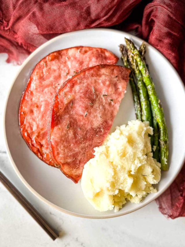 air fryer ham steak with mashed potatoes and asparagus on a white plate.