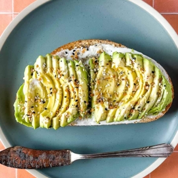 an avocado tartine sits on a blue plate with a vintage silver knife over a peach tile background.