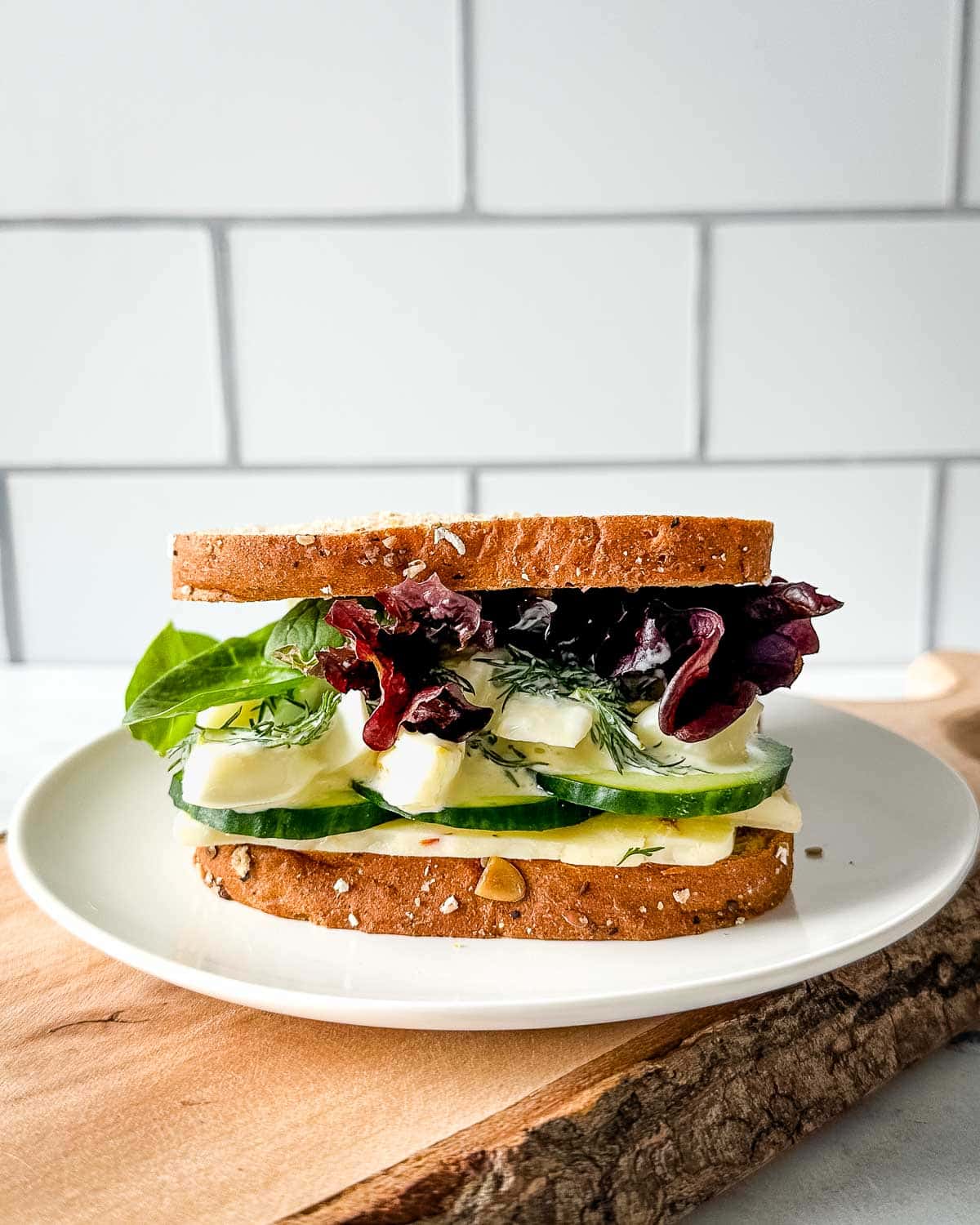 an egg and cucumber sandwich with pepper jack cheese, spring mix, and dill sauce is shown on a white plate over a rustic wooden cutting board.