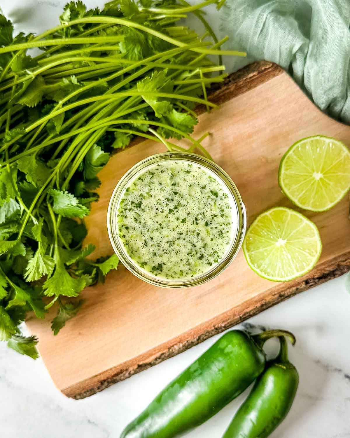 A jar of honey jalapeno lime vinaigrette sits on a cutting board surrounded by limes, cilantro, and jalapenos.