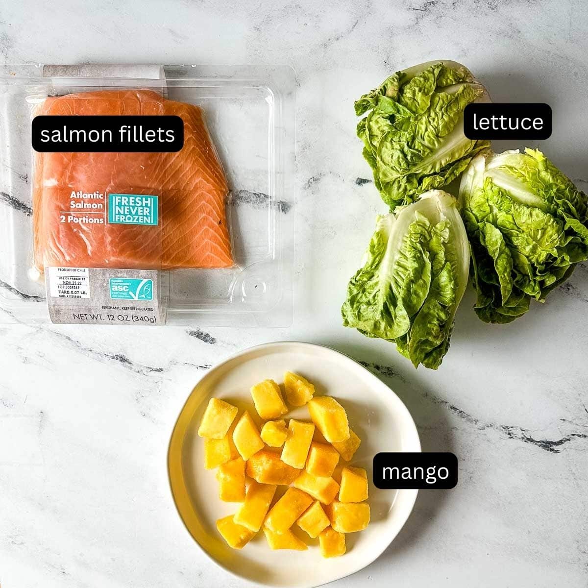 The labeled ingredients for mango salmon salad sit on a white marble counter.