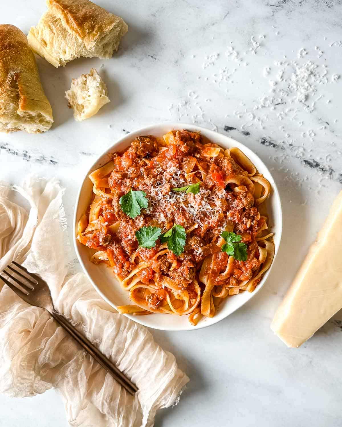 a plate of slow cooker lamb ragu over pappardelle pasta surrounded by a block of parmesan, a white linen, and torn baguette.
