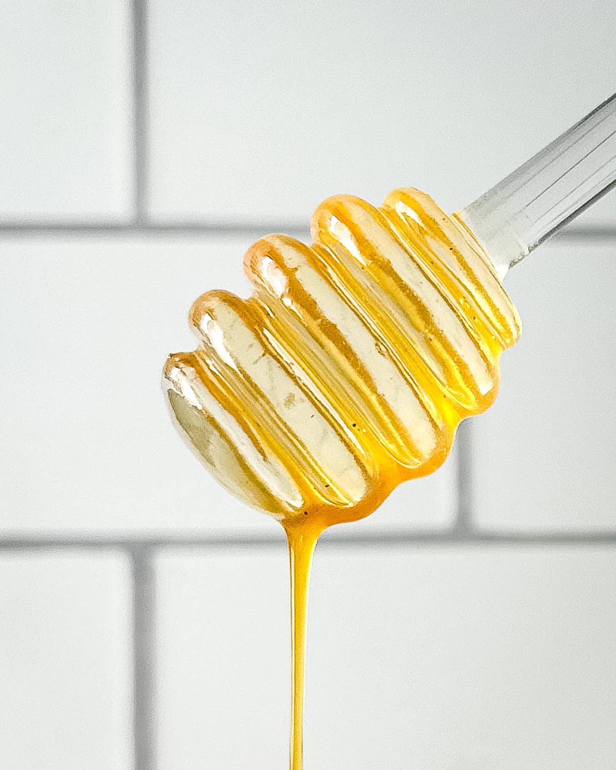 Closeup of honey dipper dripping hot honey sauce in front of a white subway tile wall.