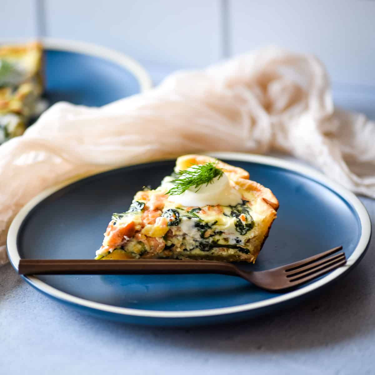 Side view of a slice of salmon spinach quiche on a dark blue plate with a copper fork.