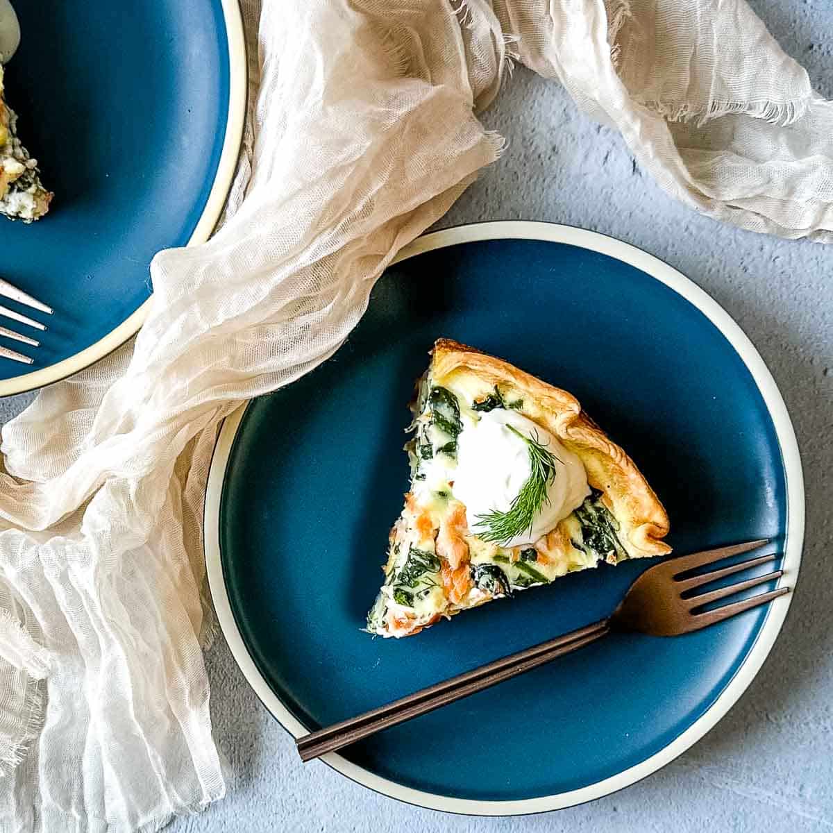 a slice of smoked salmon and spinach quiche on a teal plate with a copper fork.