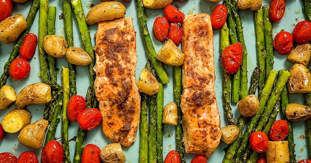 Closeup overhead shot of roasted salmon, asparagus, fingerling potatoes, and cherry tomatoes on a blue sheet tray.
