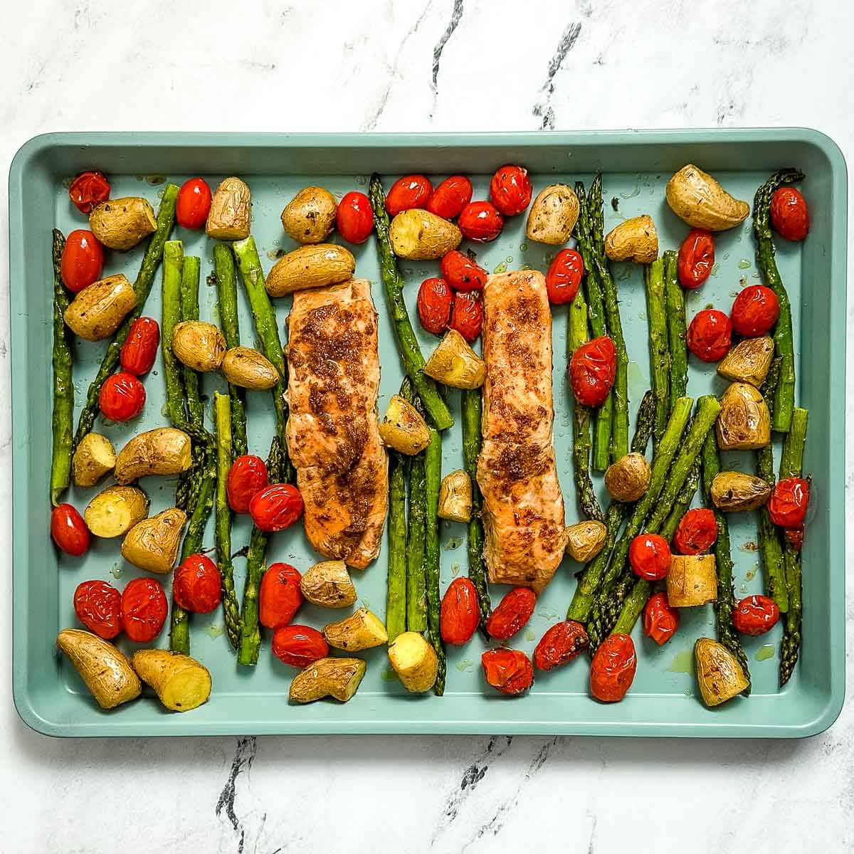 Overhead shot of roasted salmon, asparagus, fingerling potatoes, and cherry tomatoes on a blue sheet tray.