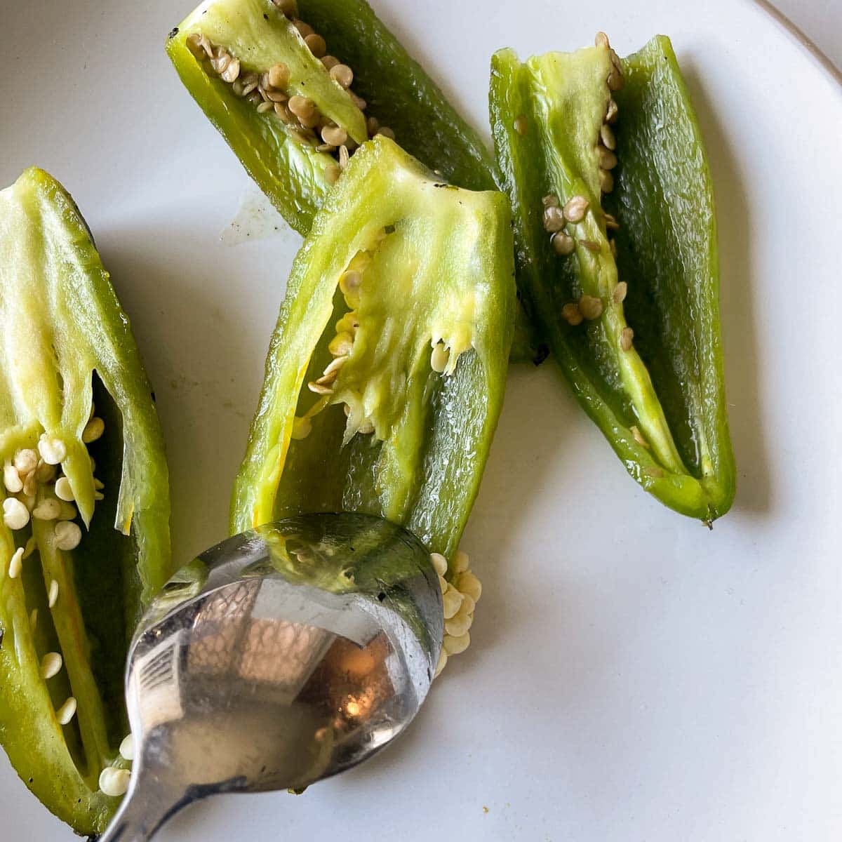 A metal spoon removes the seeds and membrane from a halved, roasted jalapeno.