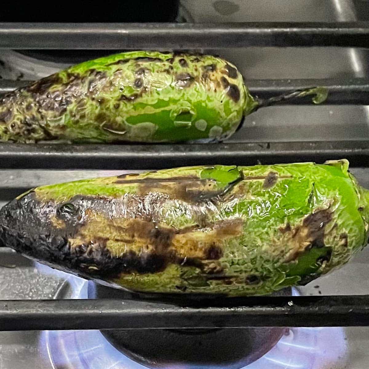 Two jalapenos are roasted over a gas flame on a stovetop.