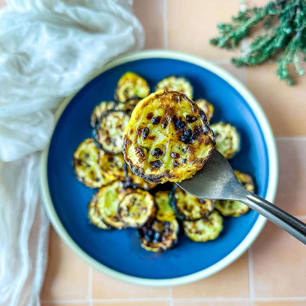 A slice of air fried summer squash is held on a fork over a plate of air fried zucchini and summer squash.