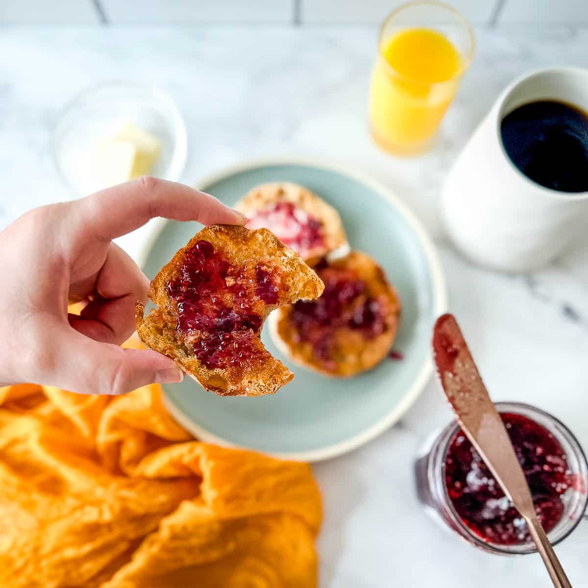 An English muffin with a bite taken out of it is held over a scene with a blue plate, a yellow linen, jam, a knife, coffee, and orange juice.