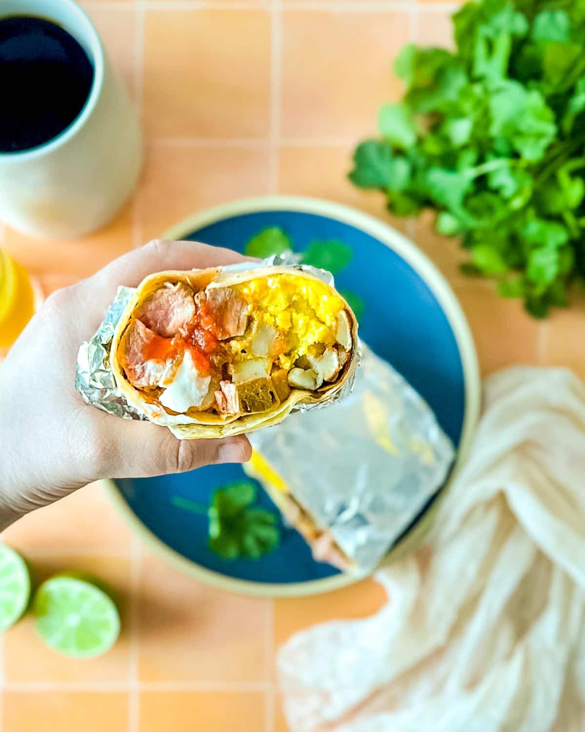 Half of a carne asada breakfast burrito is held over a blue plate that's surrounded by limes, coffee, and orange juice.