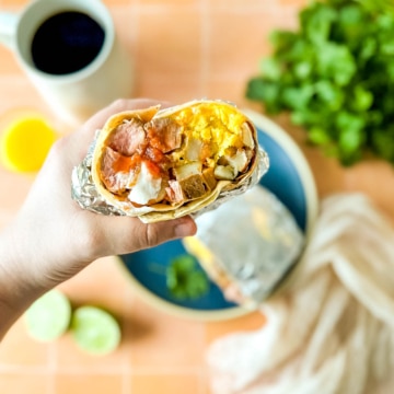 Half of a carne asada breakfast burrito is held over a blue plate surrounded by limes, coffee, orange juice, cilantro, and a white linen.