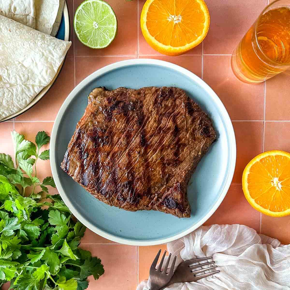 A grilled steak sits on a light blue plate surrounded by sliced oranges, limes, a bunch of cilantro, tortillas, and a glass of beer.