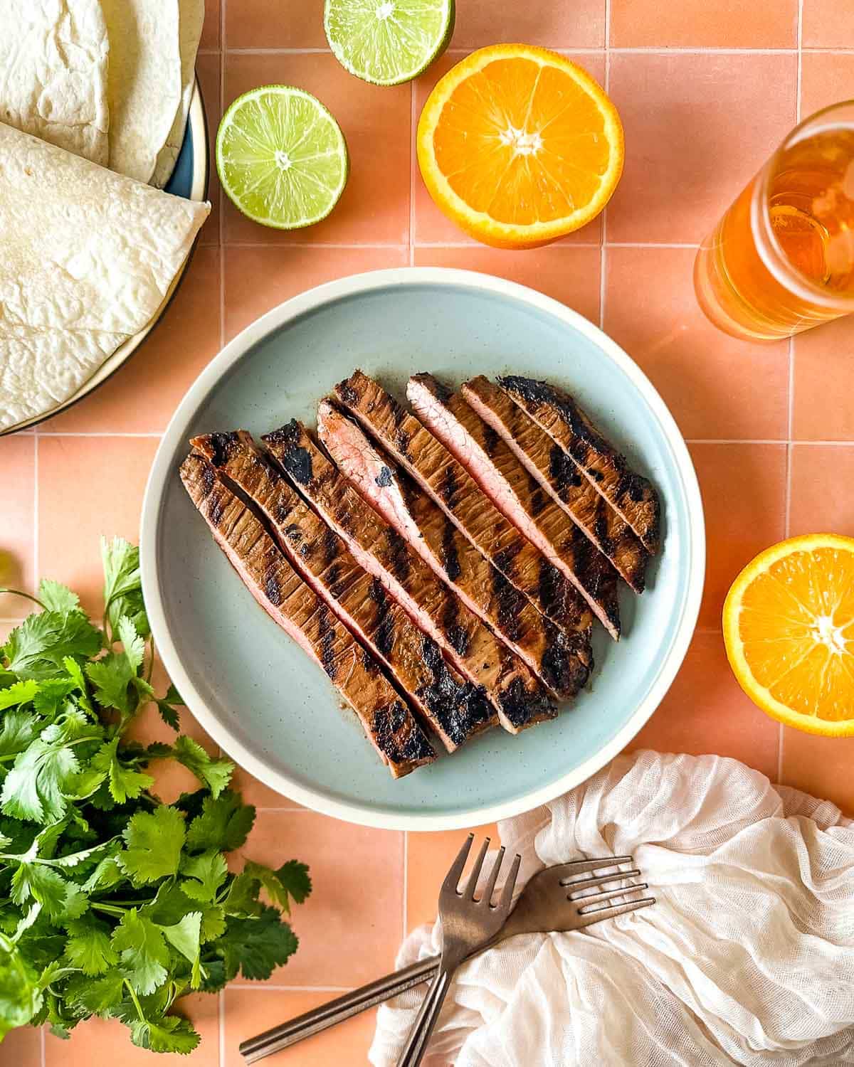 Sliced carne asada sits on a light blue plate surrounded by sliced oranges, limes, a bunch of cilantro, tortillas, and a glass of beer.