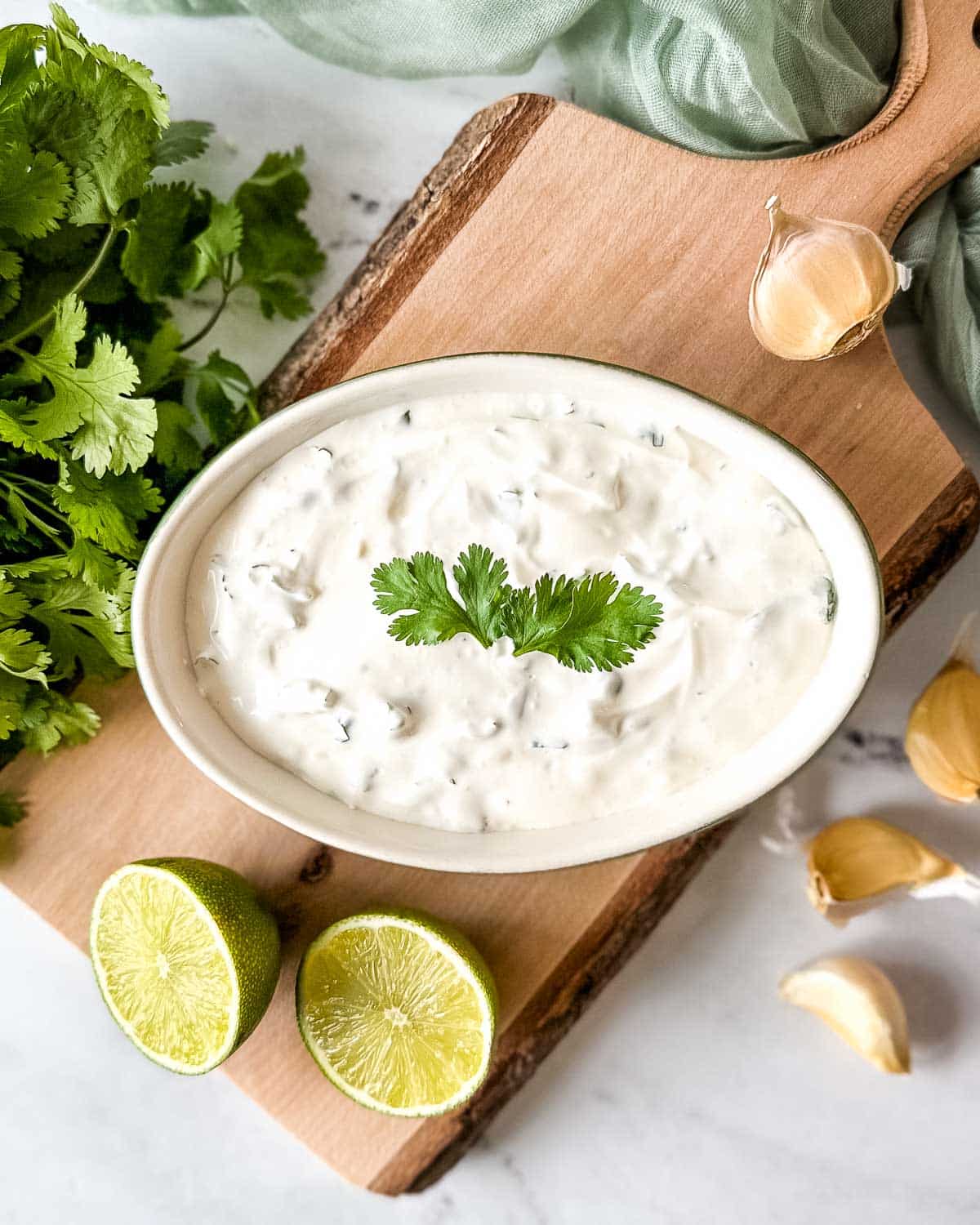 Cilantro lime crema in a white and green dish surrounded by lime halves, fresh cilantro, and garlic cloves.