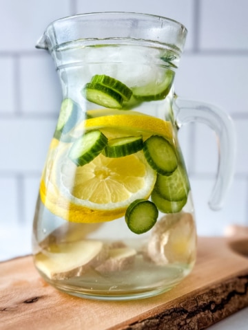A glass pitcher filled with cucumber lemon ginger water sits on a wooden cutting board.