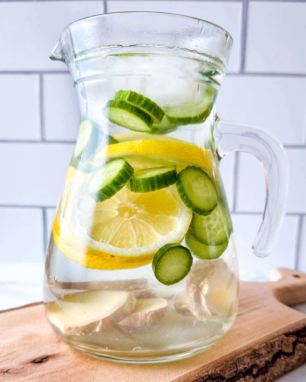 A glass pitcher filled with cucumber lemon ginger water sits on a wooden cutting board.