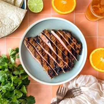 Sliced carne asada sits on a light blue plate surrounded by sliced oranges, limes, a bunch of cilantro, tortillas, and a glass of beer.