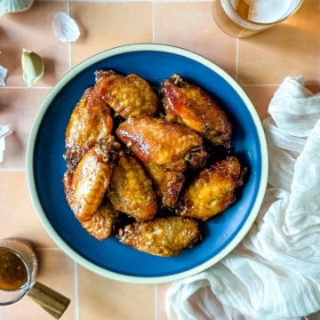 An overhead shot of soy garlic chicken wings on a blue plate surrounded by garlic cloves, a white linen, and a glass of beer.