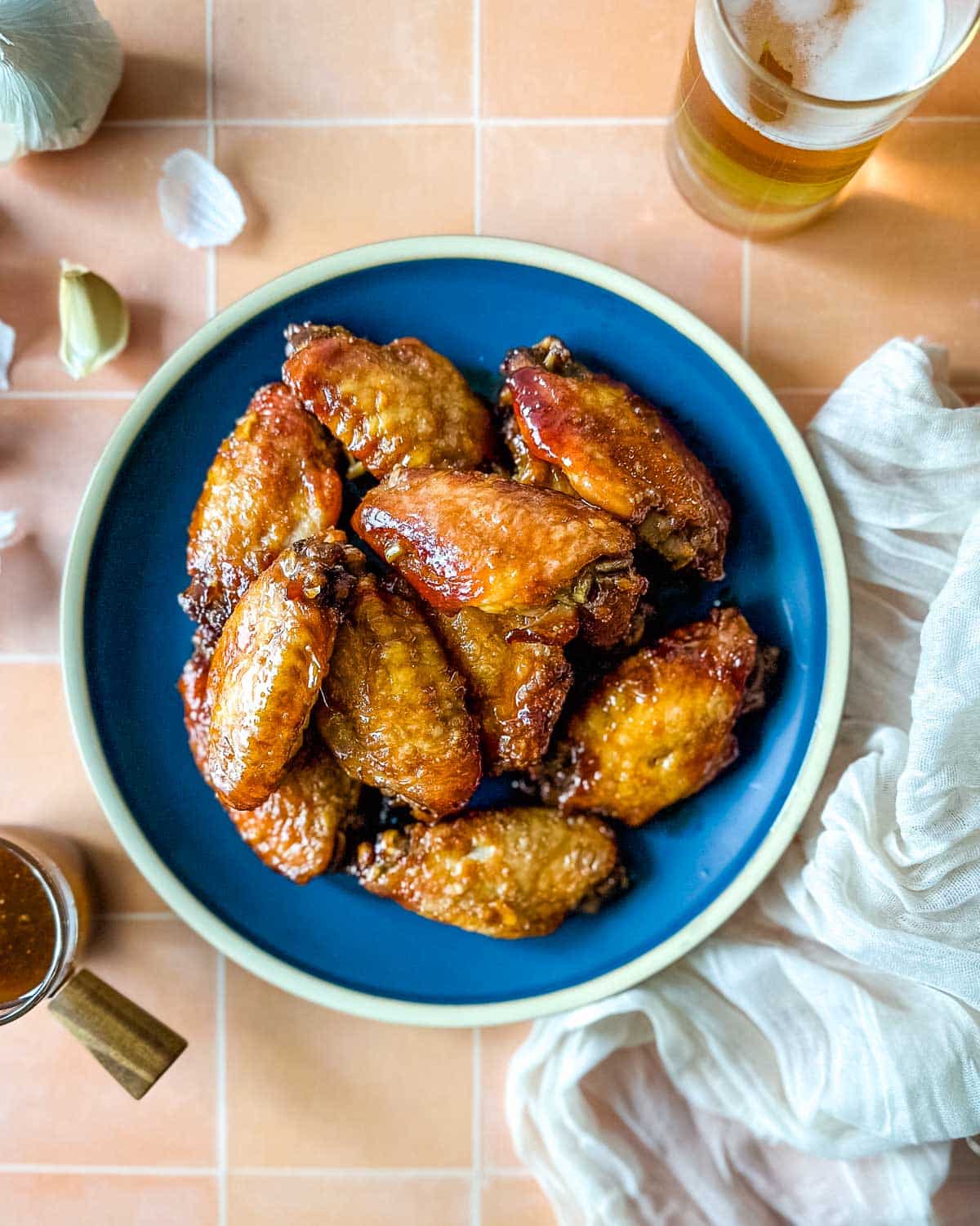 An overhead shot of soy garlic chicken wings on a blue plate surrounded by garlic cloves, a white linen, and a glass of beer.