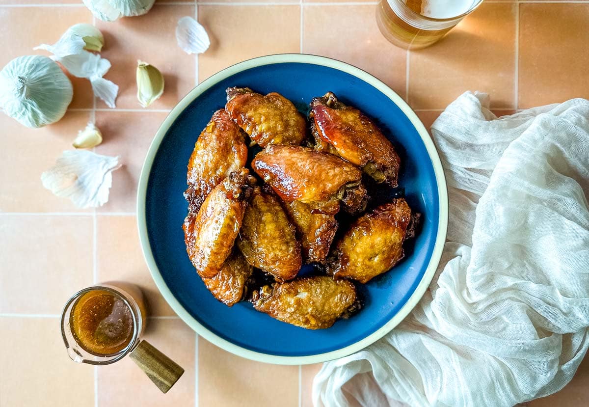 Soy garlic wings on a blue plate surrounded by a white linen, a glass of bear, soy ginger sauce, and cloves of garlic.