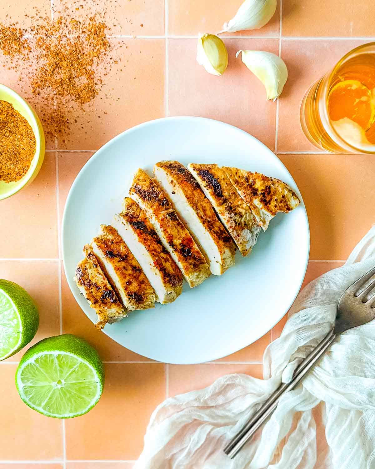 Sliced Tajín Grilled Chicken sits on a white plate surrounded by Tajín seasoning, a sliced lime, a white linen, and a small glass of beer.