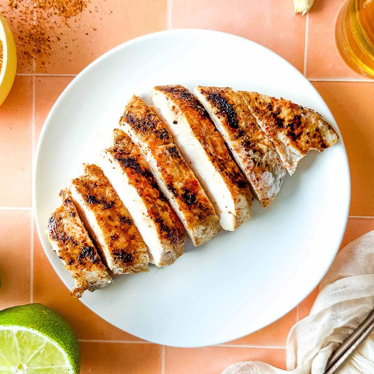 Sliced Tajín Grilled Chicken sits on a white plate surrounded by Tajín seasoning, a sliced lime, a white linen, and a small glass of beer.