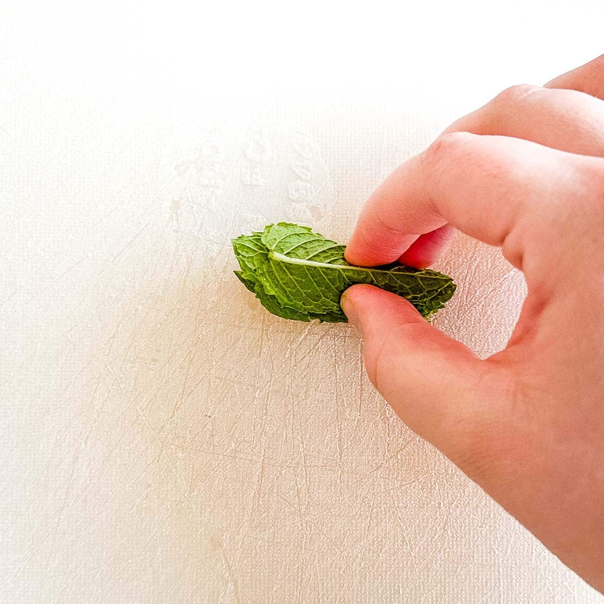 A hand holds a small pile of rolled mint leaves against a cutting board.