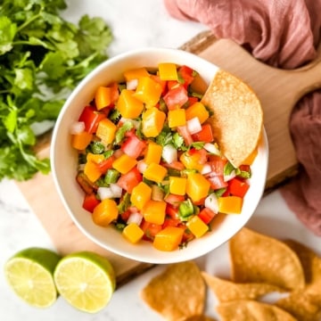 A white bowl of mango pico de gallo surrounded by tortilla chips, limes, cilantro, and a pink linen.