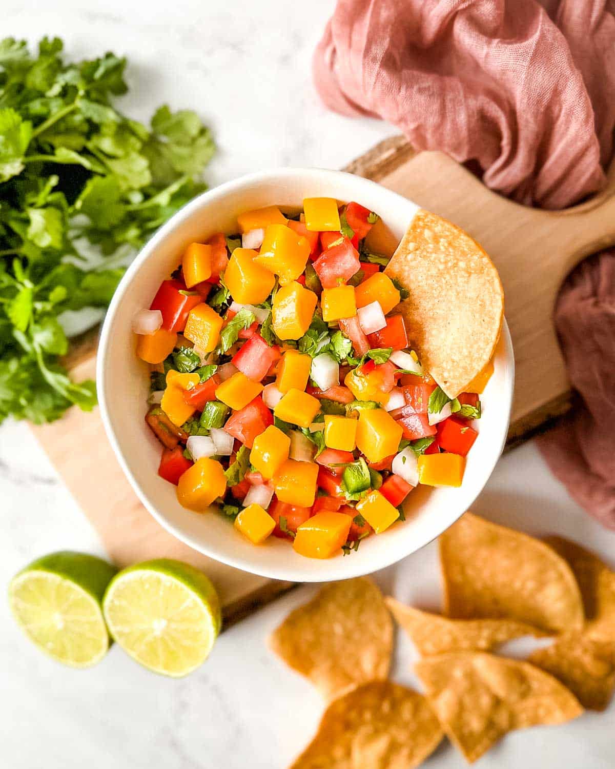 A white bowl of mango pico de gallo surrounded by tortilla chips, limes, cilantro, and a pink linen.