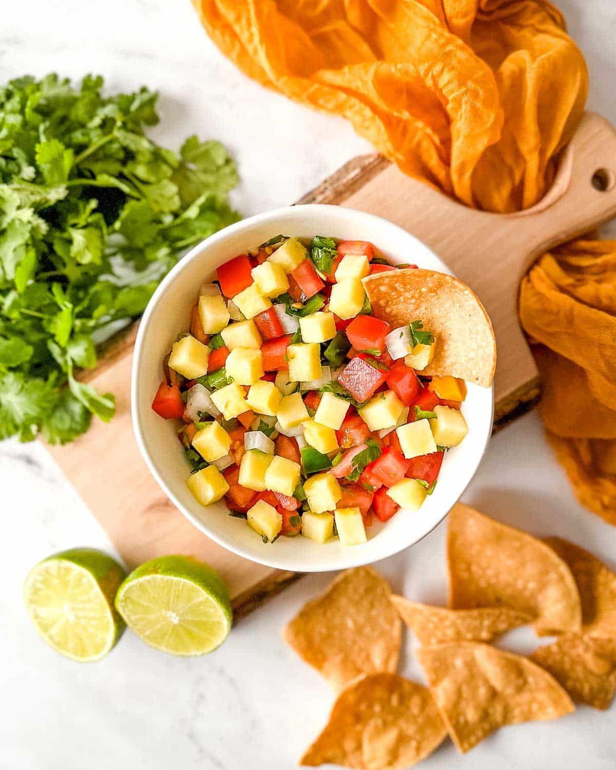 Overhead shot of pineapple pico de gallo in a white bowl surrounded by tortilla chips, a halved lime, and fresh cilantro.