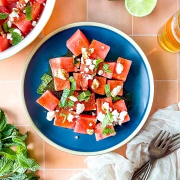 Watermelon salad garnished with mint and feta on a blue plate surrounded by cut limes, fresh mint, and a white linen.
