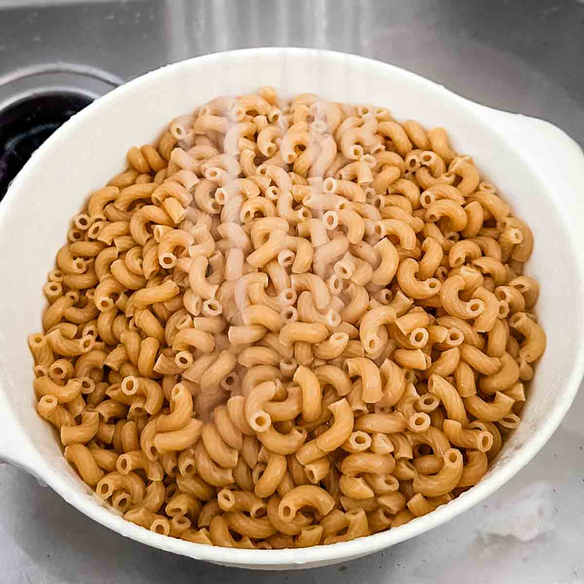 Whole wheat pasta is rinsed with cold water in a white colander.